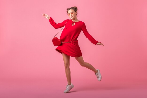 Atrakcyjna szczęśliwa uśmiechnięta stylowa kobieta w czerwonej modnej sukience skaczącej na różowej ścianie na białym tle, trend w modzie wiosna lato, dzień świętego walenity, zalotna dziewczyna