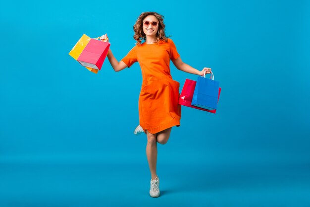 Atrakcyjna szczęśliwa uśmiechnięta stylowa kobieta zakupoholiczka w pomarańczowej modnej sukience oversize, skoki, bieganie trzymając torby na zakupy na niebieskim tle na białym tle