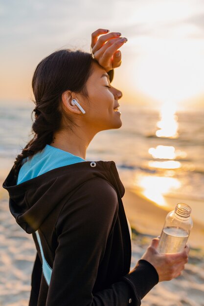 Atrakcyjna szczupła kobieta robi ćwiczenia sportowe na plaży o poranku wschód słońca w stroju sportowym, spragniona woda pitna w butelce, zdrowy tryb życia, słuchanie muzyki na bezprzewodowych słuchawkach, uśmiechanie się szczęśliwe