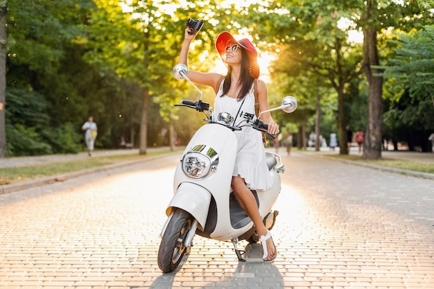 Atrakcyjna uśmiechnięta kobieta jeżdżąca na motocyklu na ulicy w letnim stroju na sobie białą sukienkę i czerwony kapelusz, podróżująca na wakacje, robienie zdjęć aparatem vintage