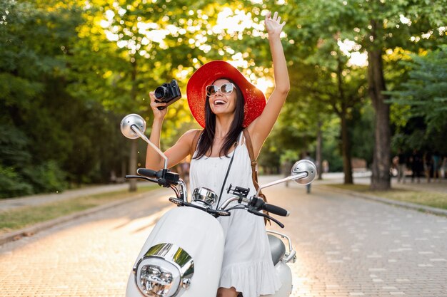 Atrakcyjna uśmiechnięta kobieta jeżdżąca na motocyklu na ulicy w letnim stylu ubrana w białą sukienkę i czerwony kapelusz, podróżująca na wakacje, robienie zdjęć aparatem vintage, machanie ręką, powitanie
