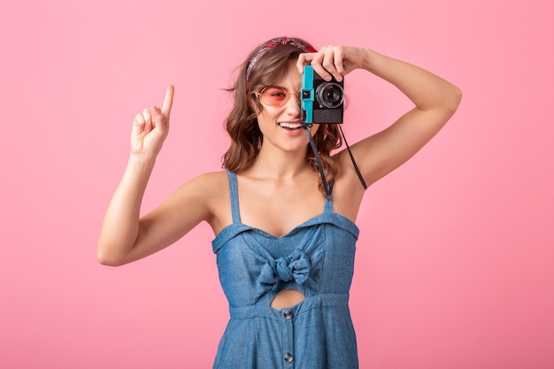 Atrakcyjna uśmiechnięta kobieta robienie zdjęć aparatem vintage, wskazując palcem w górę, ubrana w dżinsową sukienkę i okulary na białym tle na różowym tle