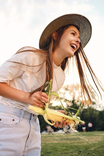 Atrakcyjna wesoła brązowowłosa dziewczyna w słomkowym kapeluszu szczęśliwie stojąca z kukurydzą na pikniku w parku miejskim
