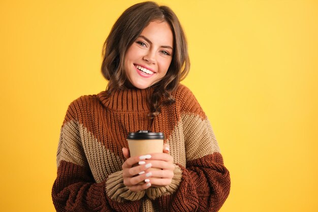 Atrakcyjna wesoła dziewczyna w przytulnym swetrze z kawą, aby iść szczęśliwie patrząc w kamerę na żółtym tle