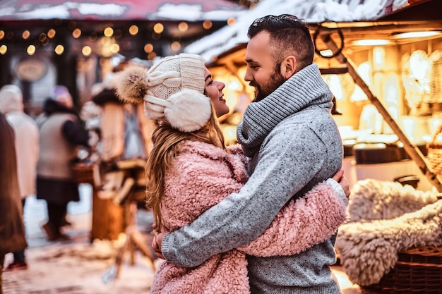 Atrakcyjna zakochana para, stylowa para w ciepłych ubraniach, przytulająca się do siebie i patrząca na siebie na zimowy jarmark w okresie świątecznym.