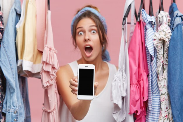 Atrakcyjna zakupoholiczka trzymająca telefon komórkowy z pustym ekranem, pokazująca szokujące ceny sprzedaży na stronie sklepu odzieżowego podczas zakupów online