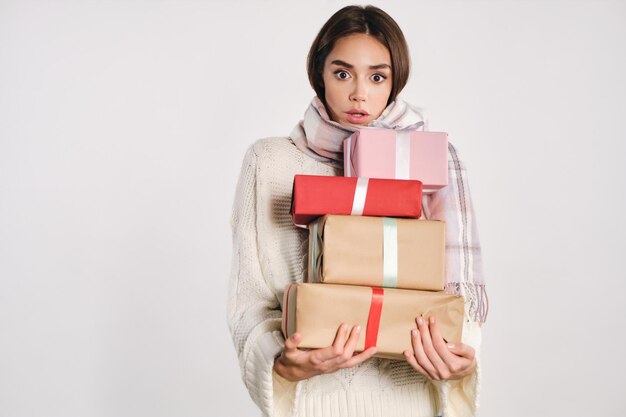 Atrakcyjna, zszokowana dziewczyna w przytulnym swetrze z szalikiem, trzymająca pudełka z prezentami, zdumiewająco patrząca w kamerę na białym tle