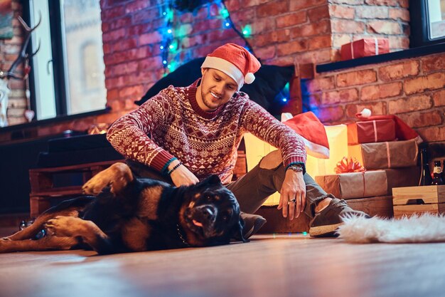 Atrakcyjny brodaty mężczyzna hipster siedzi na podłodze ze swoim psem Rottweiler w pokoju z dekoracją świąteczną.