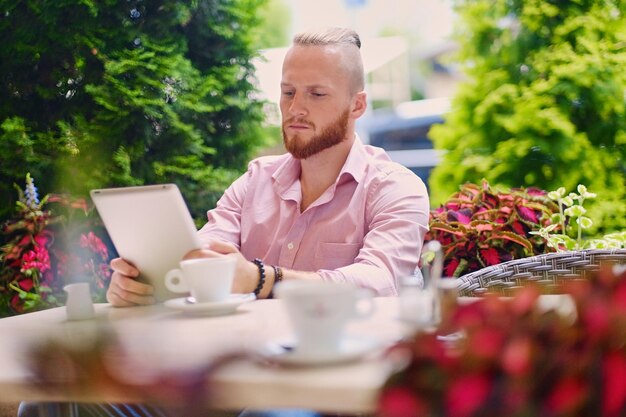 Atrakcyjny brodaty mężczyzna w różowej koszuli siedzi przy stoliku w kawiarni i korzysta z komputera typu tablet.
