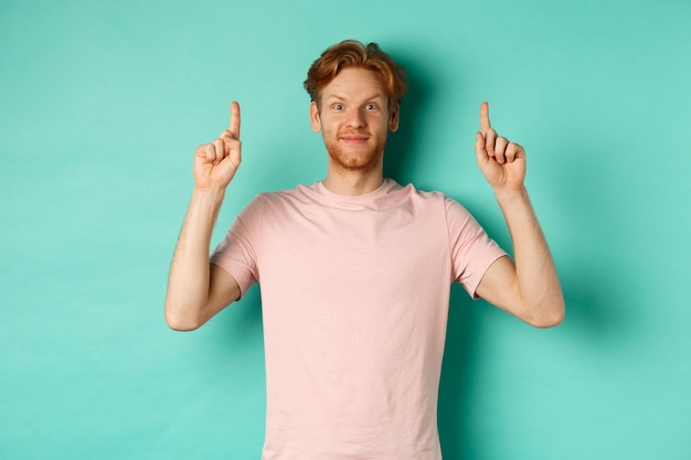 Bezpłatne zdjęcie atrakcyjny brodaty mężczyzna z rudymi włosami, ubrany w t-shirt, uśmiechnięty wesoło i wskazujący palcem w górę, pokazujący reklamę, stojący nad turkusowym tłem.