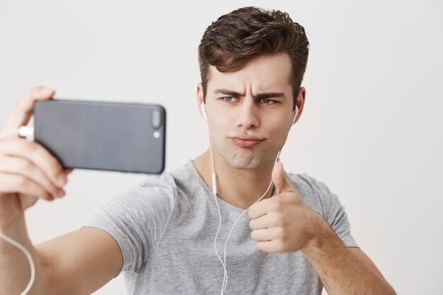 Atrakcyjny kaukaski mężczyzna o ciemnych włosach, marszcząc brwi, trzymając telefon komórkowy, trzymając kciuk do góry, jednocześnie stwarzając dla selfie. Ludzka mimika, emocje i gesty.
