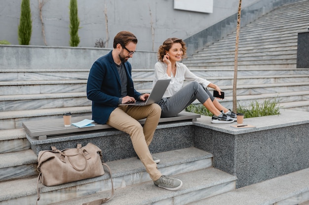 Atrakcyjny mężczyzna i kobieta siedzący na schodach w centrum miasta, rozmawiający przez bezprzewodowe słuchawki głośnomówiące