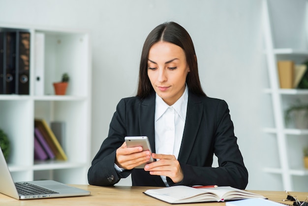 Atrakcyjny młody bizneswoman sprawdza telefon komórkowego przy miejscem pracy