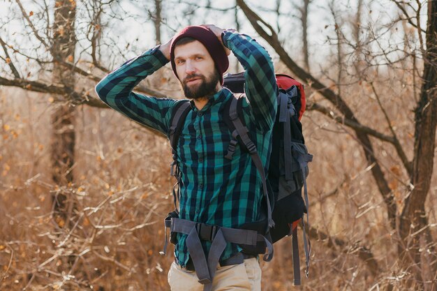 Atrakcyjny młody hipster mężczyzna podróżujący z plecakiem w lesie jesienią na sobie koszulę w kratę i kapelusz, aktywny turysta, odkrywanie przyrody w zimnych porach roku