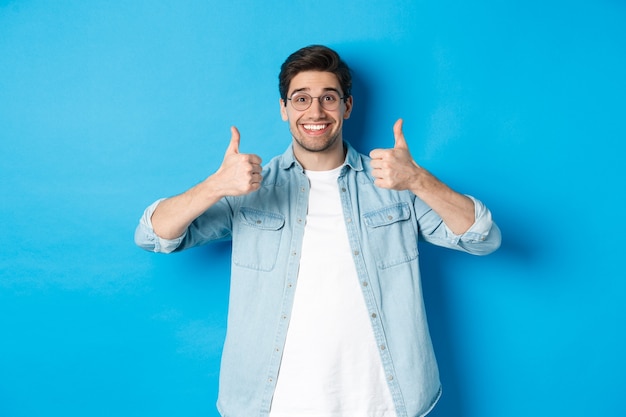 Atrakcyjny młody mężczyzna w okularach i ubraniu, pokazując kciuk do góry w aprobacie, jak coś, stojąc na niebieskim tle.