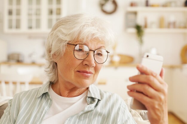 Atrakcyjny nowoczesny starszy żeński emeryt w okrągłe okulary siedzi na kanapie, trzymając ogólny telefon komórkowy, czytając sms. Emerytowana siwowłosa kobieta przeglądająca internet przy użyciu połączenia bezprzewodowego 4 g