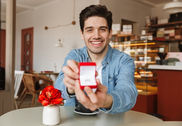 Atrakcyjny uśmiechnięty mężczyzna siedzi przy stole w restauracji i proponuje swojej kobiecie, dając pudełko z pierścionkiem zaręczynowym