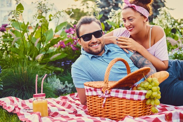 Atrakcyjny wytatuowany, brodaty mężczyzna i ruda kobieta leżące na kocu na trawniku w parku. Czas na piknik.