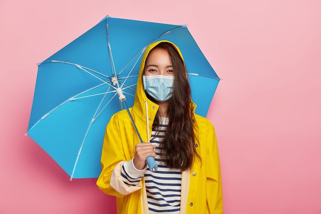 Azjatka nosi maskę ochronną, stawia czoła zanieczyszczeniu powietrza w deszczowy dzień, stoi pod parasolem, ubrana w żółty płaszcz przeciwdeszczowy