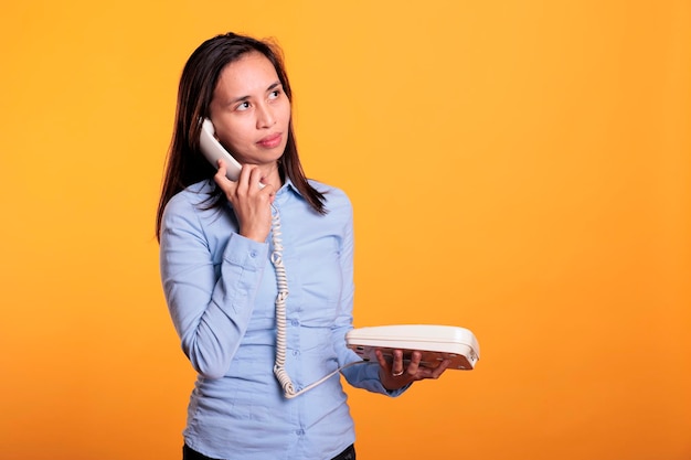 Bezpłatne zdjęcie azjatka rozmawia przez telefon stacjonarny ze zdalnym pracownikiem, rozmawia na linii telefonicznej w stylu retro z kablem w studiu na żółtym tle. wesoły, uśmiechnięty młody dorosły, cieszący się dyskusją o stylu życia