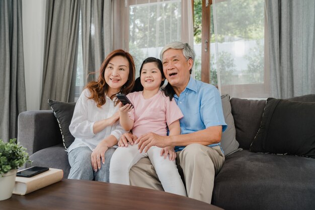 Azjatyccy dziadkowie oglądają telewizję z wnuczką w domu. Starszy chińczyka, dziadka i babci szczęśliwy używa rodzinny czas, relaksuje z młoda dziewczyna dzieciaka lying on the beach na kanapie w żywym izbowym pojęciu.