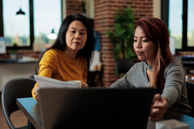 Azjatyccy przedsiębiorcy pracujący przy projekcie marketingowym analizującym statystyki dotyczące laptopa, przeprowadzający burze mózgów podczas spotkania biznesowego w startupowym biurze. Współpracujący menedżerowie wykonawczy