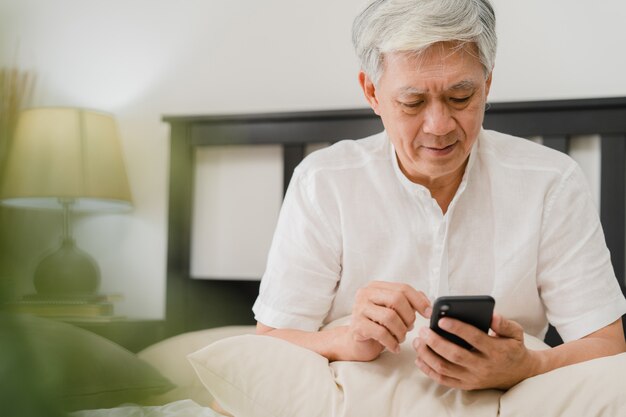 Azjatyccy starsi mężczyzna używa telefon komórkowego w domu. Azjatycka Starsza Chińska męska rewizi informacja o dlaczego dobre zdrowie na internecie podczas gdy kłamający na łóżku w sypialni w domu w ranku pojęciu.