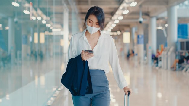 azjatycka biznesowa dziewczyna za pomocą smartfona do czekania karty pokładowej, chodzenie z bagażem do terminala na locie krajowym na lotnisku.