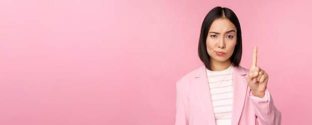 Azjatycka bizneswoman z poważnym zaniepokojonym wyrazem twarzy pokazującym tabu zatrzymania ruchu zakaz gestu potępienia czegoś złego stojącego na różowym tle