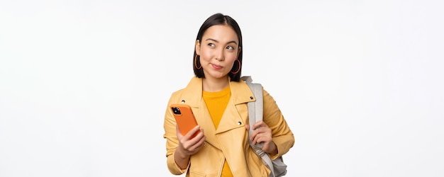 Azjatycka dziewczyna podróżująca z plecakiem trzymająca telefon komórkowy za pomocą aplikacji na smartfona, wyglądająca na zamyśloną stojącą na białym tle