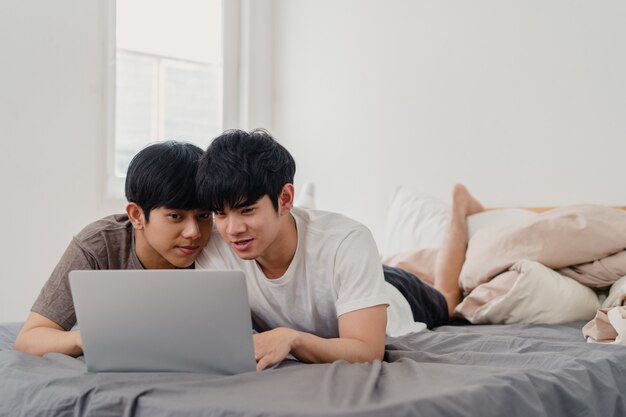 Azjatycka Homoseksualna lgbtq mężczyzna para używa komputerowego laptop w nowożytnym domu. Młody miłośnik Azji mężczyzna szczęśliwy zrelaksować się odpocząć po przebudzeniu, oglądając film leżący na łóżku w sypialni w domu rano.