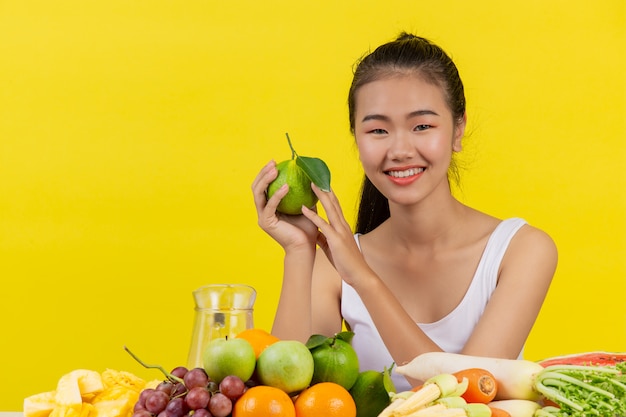 Azjatycka kobieta jest ubranym białego podkoszulek bez rękawów. Trzymanie pomarańczy prawą ręką Na stole jest wiele różnych owoców.