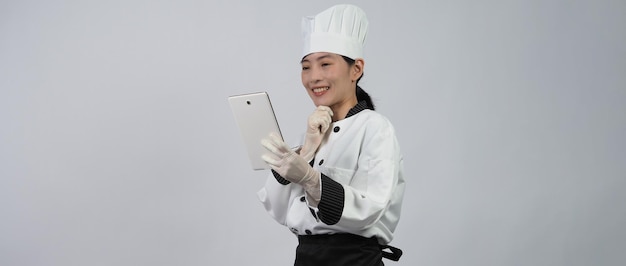 Azjatycka kobieta kucharz trzyma smartfon lub tablet cyfrowy i otrzymała zamówienie ze sklepu internetowego lub aplikacji handlowej. ona uśmiecha się w mundurze szefa kuchni i stojąc na białym tle. sprzedawca żywności online.
