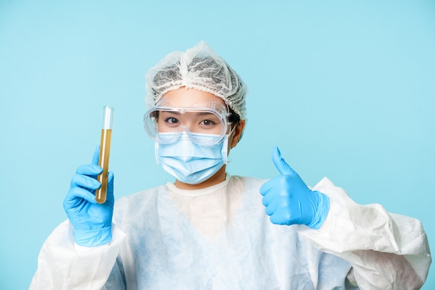 Azjatycka lekarka, pracownica laboratorium w osobistym sprzęcie ochronnym, pokazująca kciuki do góry i analizująca probówkę z próbką, stojąca na niebieskim tle