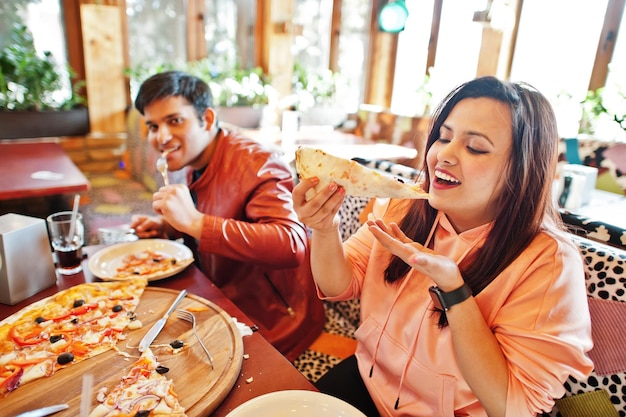 Azjatycka para przyjaciół jedząca pizzę podczas imprezy w pizzerii Szczęśliwi ludzie z Indii bawią się razem jedząc włoskie jedzenie i siedząc na kanapie