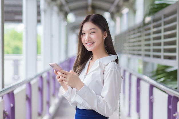 Azjatycka piękna biznesowa kobieta stoi na estakadzie kolejki miejskiej w mieście i korzysta ze swojego smartfona