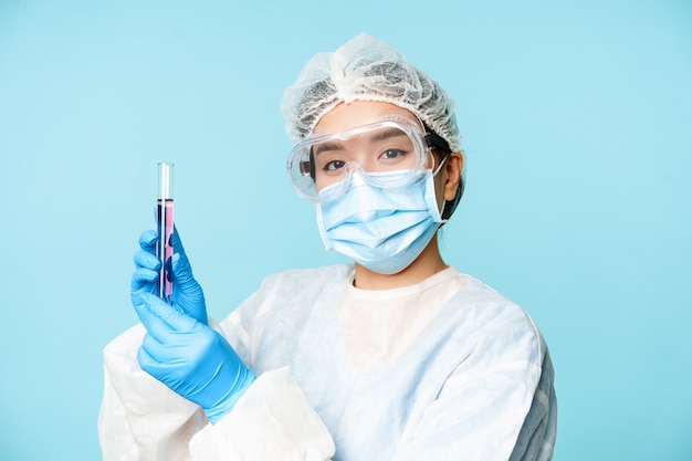 Azjatycka pielęgniarka lub pracownik laboratorium w osobistym sprzęcie ochronnym pokazująca probówkę z próbką stojącą w medi...