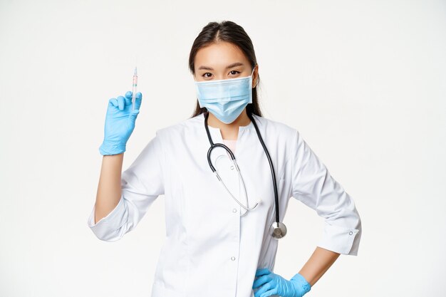 Azjatycka pielęgniarka w masce medycznej trzymająca strzykawkę ze szczepionką, pojęcie opieki zdrowotnej i szczepień, stojąca w mundurze kliniki, białe tło.
