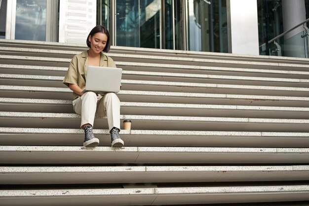 Azjatycka studentka siedzi na schodach w pobliżu kampusu na laptopie, odrabia pracę domową na świeżym powietrzu