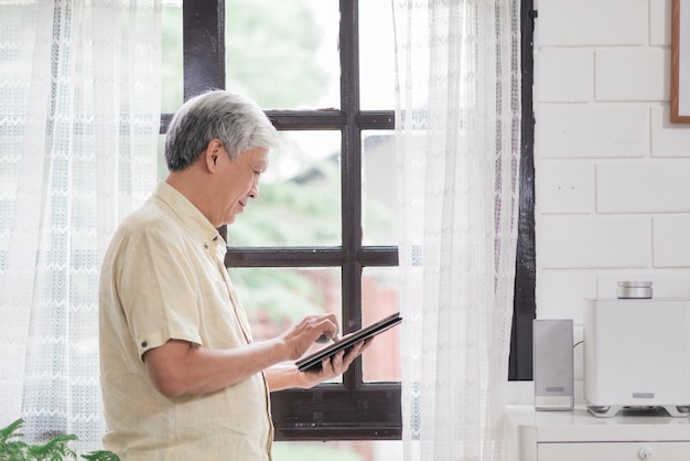 Azjatycki starsza osoba mężczyzna używa pastylkę sprawdza ogólnospołecznego medialnego pobliskiego okno w żywym pokoju w domu. Stylu życia starszych mężczyzna w domu pojęcie.