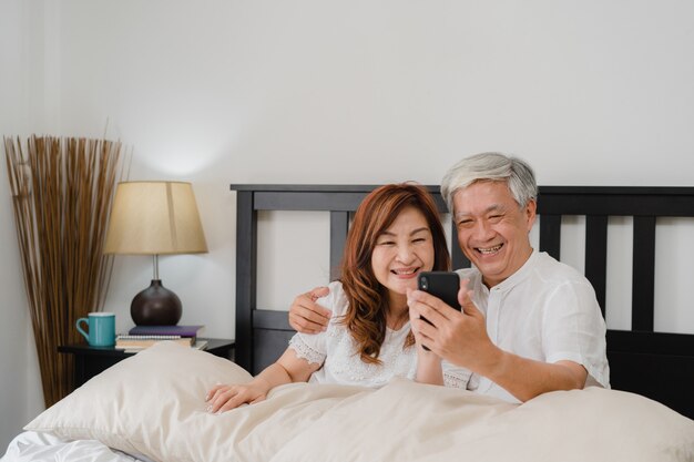 Azjatycki starszy pary selfie w domu. Azjatyccy Starsi Chińscy dziadkowie, mąż i żona szczęśliwi używa telefonu komórkowego selfie po, budzili się kłamać na łóżku w sypialni w domu w ranku pojęciu.