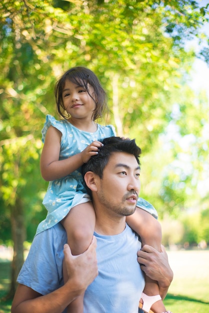 Azjatycki tata i małe dziecko odpoczynek w parku miejskim. Szczęśliwy człowiek spędza letni dzień w parku spacerując i niosąc swoją piękną córkę na ramionach. Koncepcja spędzania czasu wolnego i rodzicielstwa