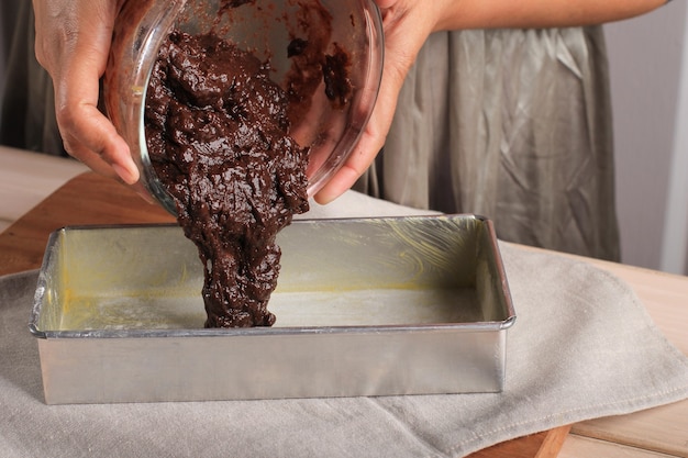 Baker ręce wlewając ciasto czekoladowe (mieszanka) z miski do wstępnie przygotowanej patelni do pieczenia. koncepcja składników i metody gotowania na białym drewnianym tle, przepisy na desery i domowe jedzenie
