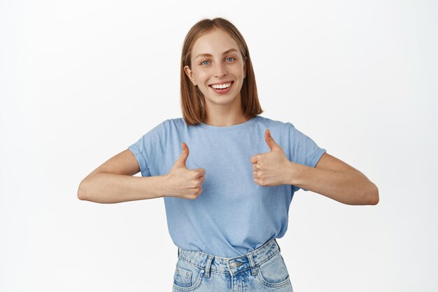 Bardzo dobrze. Atrakcyjna młoda kobieta w niebieskiej koszulce, pokazująca kciuki do góry i uśmiechnięta zadowolona, zadowolona, chwali dobrą pracę, komplementuje, zatwierdza i zgadza się, jak to, stojąc na białym tle.