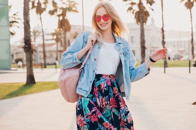 Bardzo uśmiechnięta kobieta spacerująca ulicą miasta w stylowej drukowanej spódnicy i dżinsowej kurtce oversize w różowych okularach przeciwsłonecznych, trend w letnim stylu