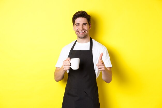 Barista przynoszący kawę i wskazujący palcem pistolet na aparat, stojący w czarnym fartuchu na żółtym tle.