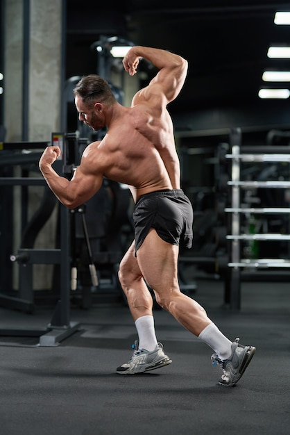 Bezpłatne zdjęcie bez koszuli kaukazyjski kulturysta demonstrujący muskularne ciało w siłowni pełna długość atrakcyjnego skoncentrowanego