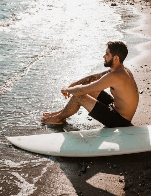 Bezpłatne zdjęcie bez koszuli mężczyzna z surfboard relaksuje blisko falowania morza