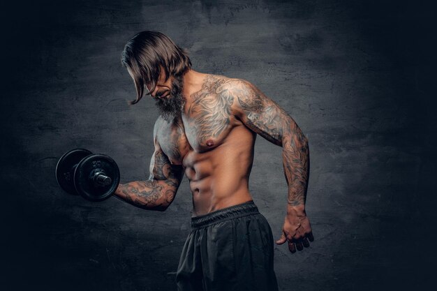 Bez koszuli, muskularny, brodaty mężczyzna z tatuażem na torsie, ćwiczący biceps z hantlami na ciemnoszarym tle.