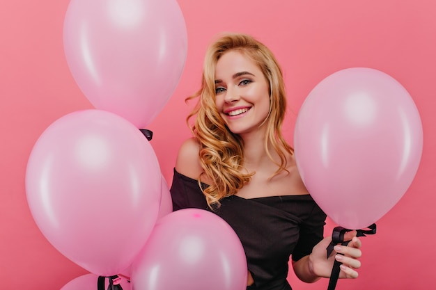 Beztroska biała dziewczyna ze szczerym uśmiechem pozuje w pobliżu różowych balonów. Wewnątrz zdjęcie przyjemnej młodej kobiety z falowanymi włosami, świętującej urodziny.